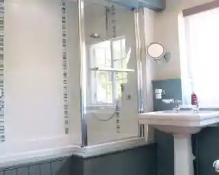 A modern bathroom featuring a glass shower enclosure, a freestanding bathtub, and a white sink. The walls are decorated with light tiles and blue accents. Sunlight filters in through a window.