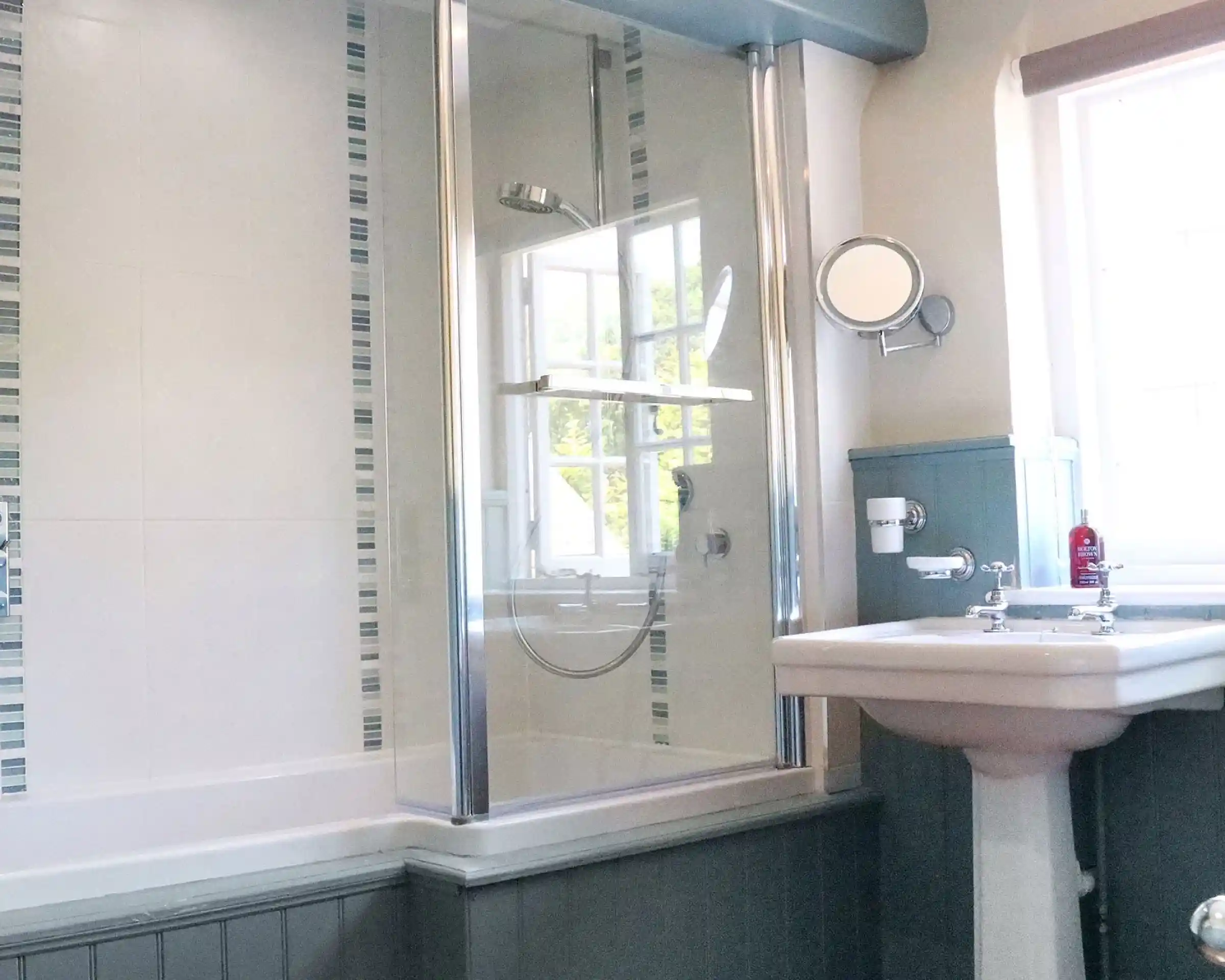 A modern bathroom featuring a glass shower enclosure, a freestanding bathtub, and a white sink. The walls are decorated with light tiles and blue accents. Sunlight filters in through a window.