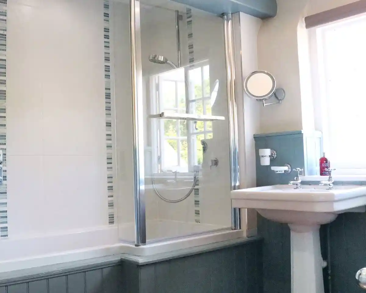 A modern bathroom featuring a glass shower enclosure, a freestanding bathtub, and a white sink. The walls are decorated with light tiles and blue accents. Sunlight filters in through a window.