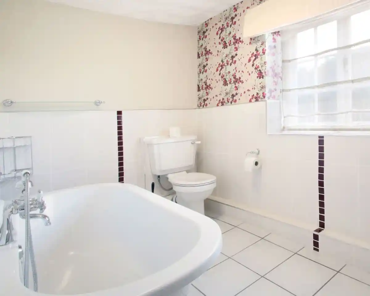 A bright bathroom featuring a white bathtub, a toilet, and tiled floors. One wall is decorated with floral wallpaper while the others have white tiles with maroon accents. A light window treatment allows natural light to fill the space.