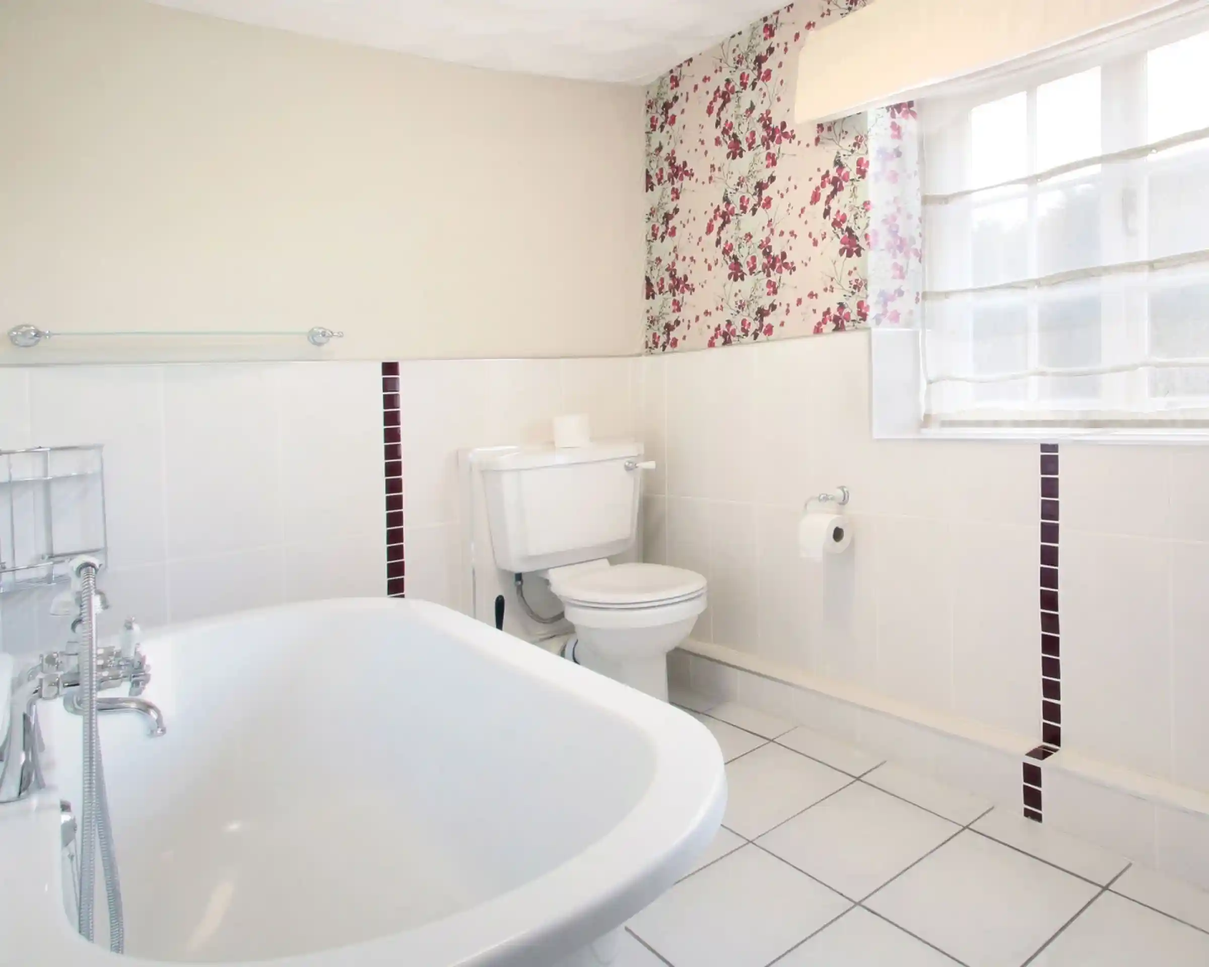 A bright bathroom featuring a white bathtub, a toilet, and tiled floors. One wall is decorated with floral wallpaper while the others have white tiles with maroon accents. A light window treatment allows natural light to fill the space.