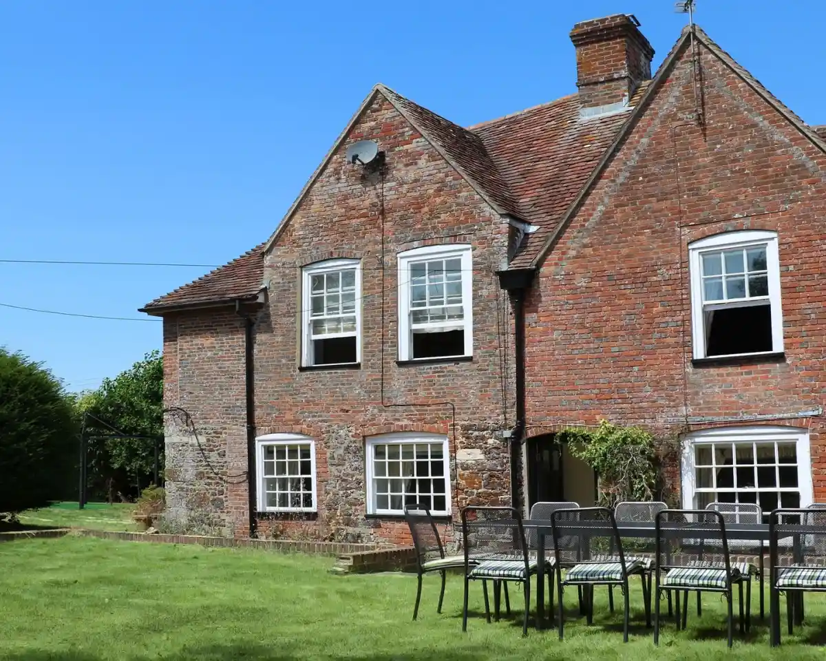 A charming brick house with multiple windows and a red roof sits in a green yard. Several black chairs are arranged on the grass in the foreground, under a clear blue sky.
