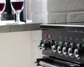 A close-up of a black kitchen range with shiny knobs. Two elegant glasses of red liquid are visible in the background on a countertop.