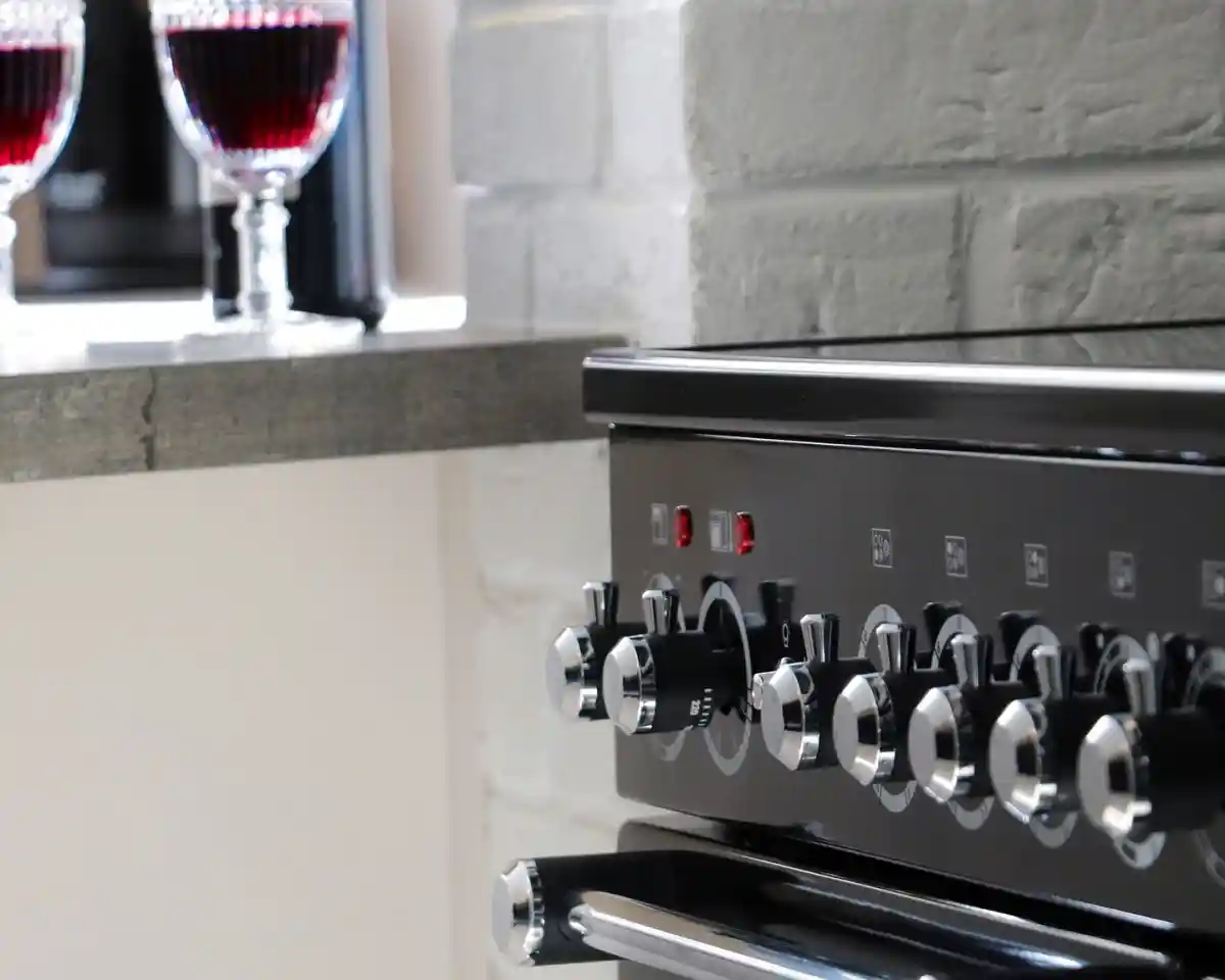 A close-up of a black kitchen range with shiny knobs. Two elegant glasses of red liquid are visible in the background on a countertop.