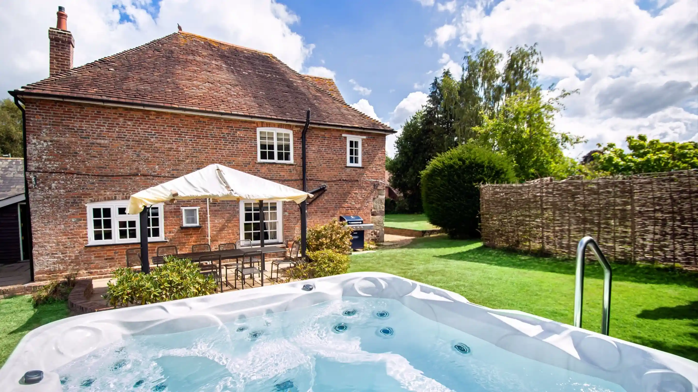 A backyard scene featuring a hot tub in the foreground, with a brick house and a patio area with outdoor seating in the background. Lush green grass surrounds the space, and trees and a fence add privacy. The sky is partly cloudy.