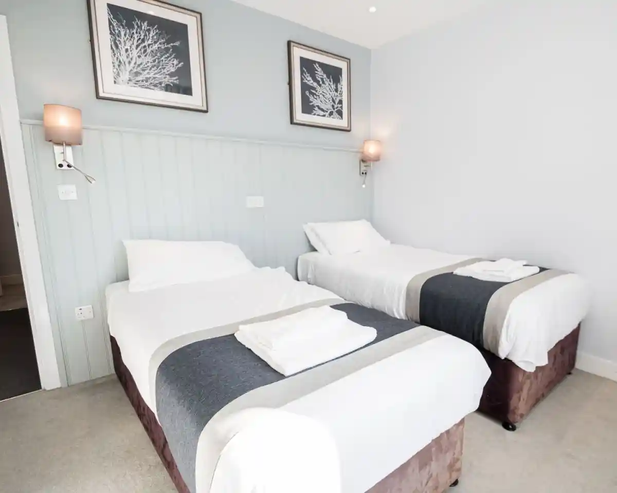 A modern hotel room featuring two single beds with white linens and gray accents. The walls are light blue with framed artwork. Wall-mounted lamps provide soft lighting.