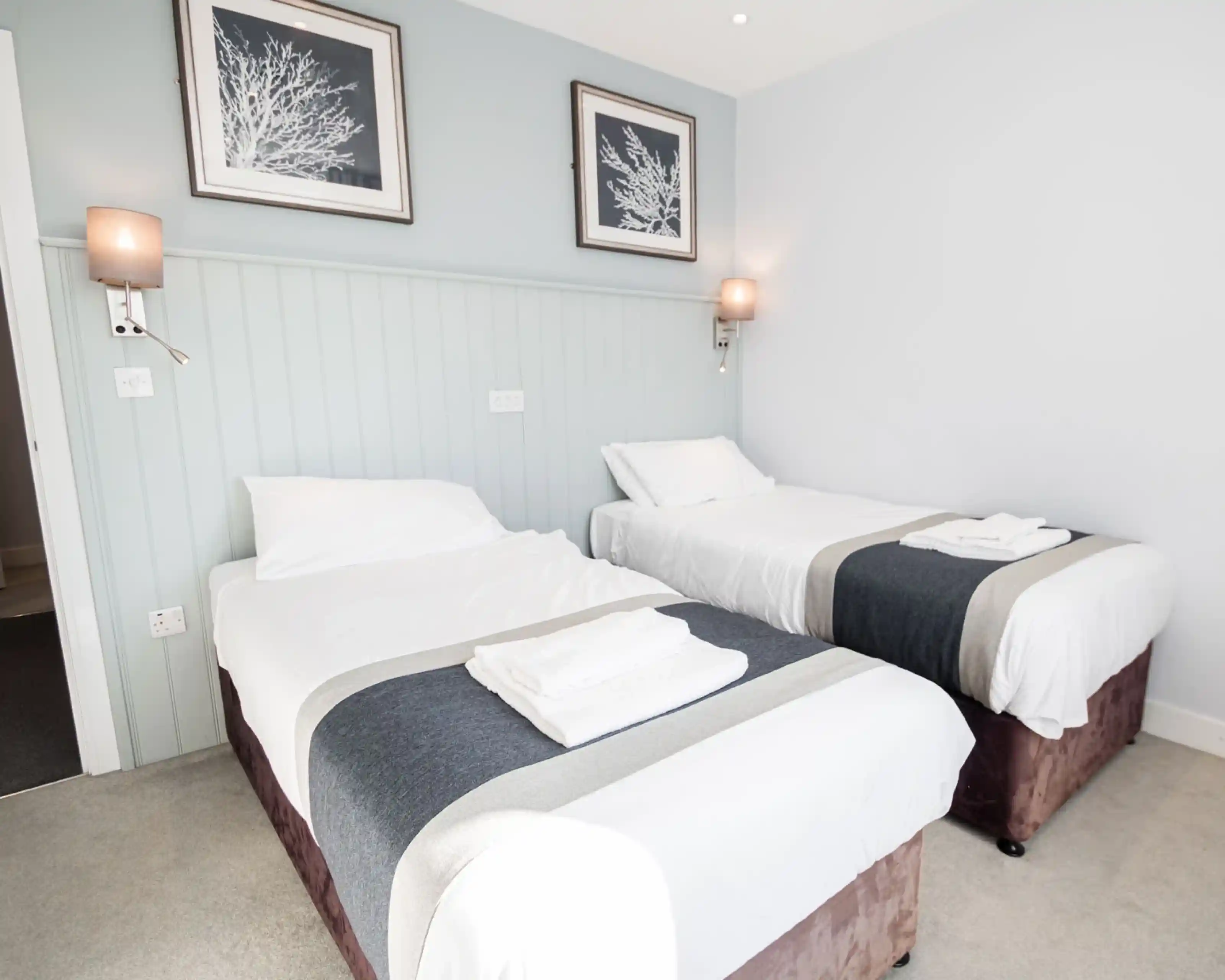 A modern hotel room featuring two single beds with white linens and gray accents. The walls are light blue with framed artwork. Wall-mounted lamps provide soft lighting.