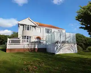 A large, two-story house with a light gray exterior and a red-tiled roof. It features a wrap-around porch and a glass-enclosed area at the back. The property is surrounded by a lush green lawn and trees under a clear blue sky.