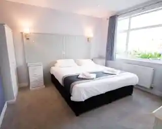 A cozy bedroom featuring a large double bed with white linens and gray accents. There are two bedside tables with lamps, a white wardrobe, and a window allowing natural light to fill the room. Soft carpet covers the floor.
