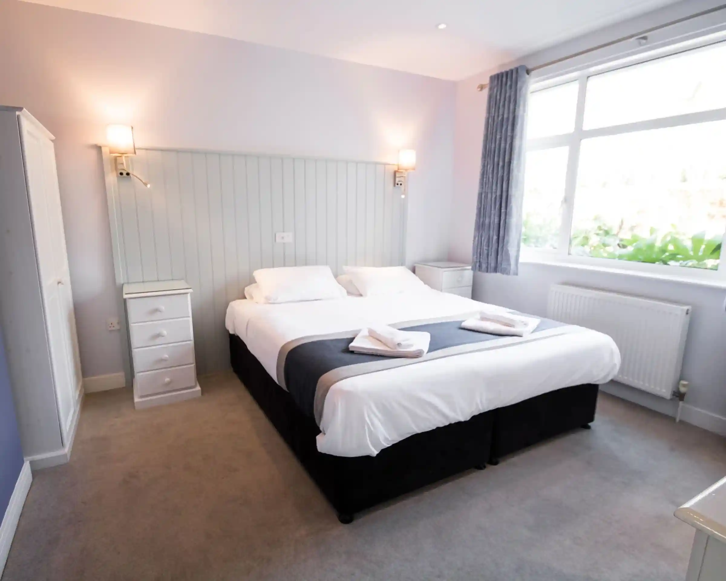 A cozy bedroom featuring a large double bed with white linens and gray accents. There are two bedside tables with lamps, a white wardrobe, and a window allowing natural light to fill the room. Soft carpet covers the floor.