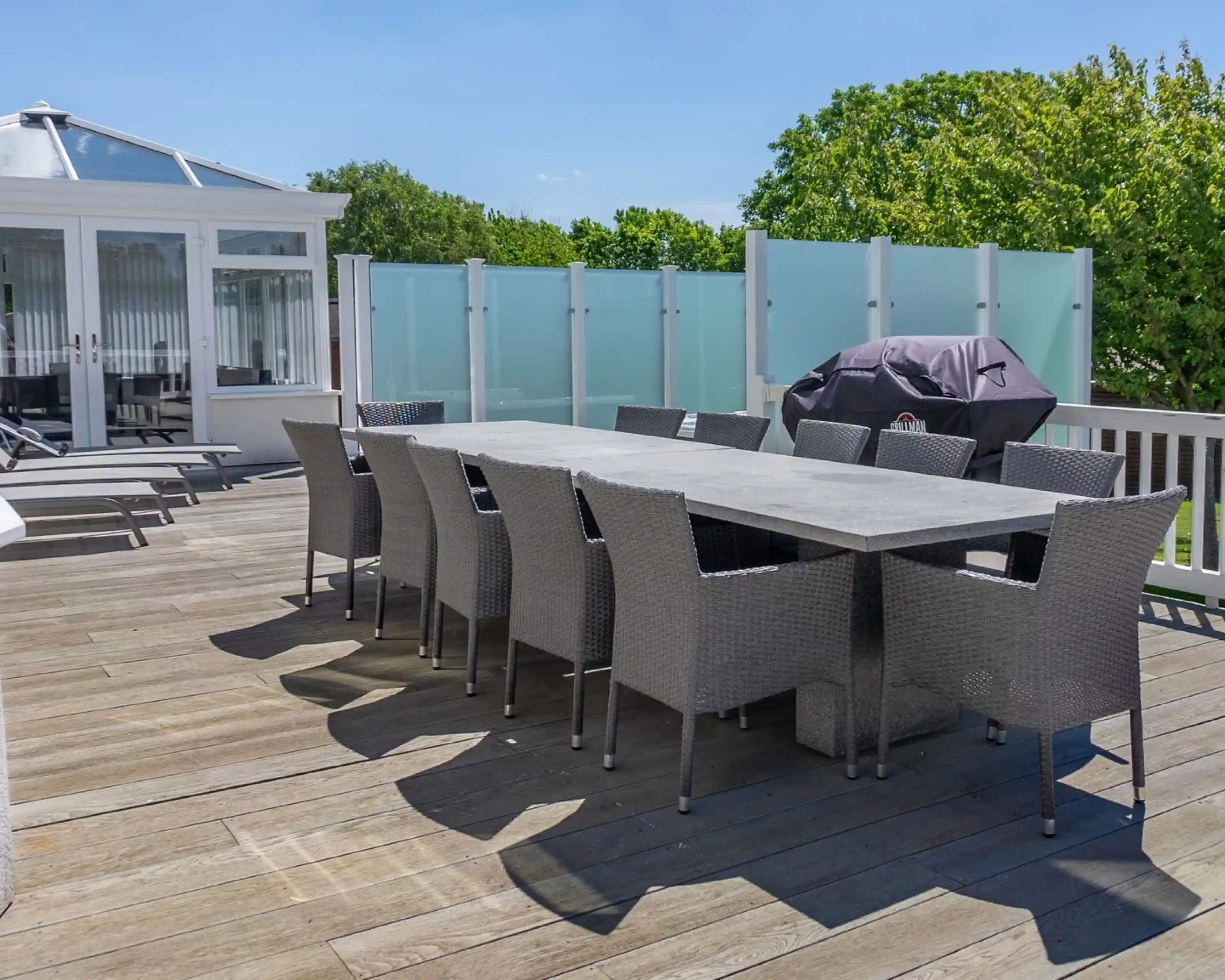 A large outdoor dining table with multiple wicker chairs is set on a wooden deck, surrounded by green trees. A grill is covered near the table, and a glass partition provides privacy. Bright blue sky with a few clouds is visible.