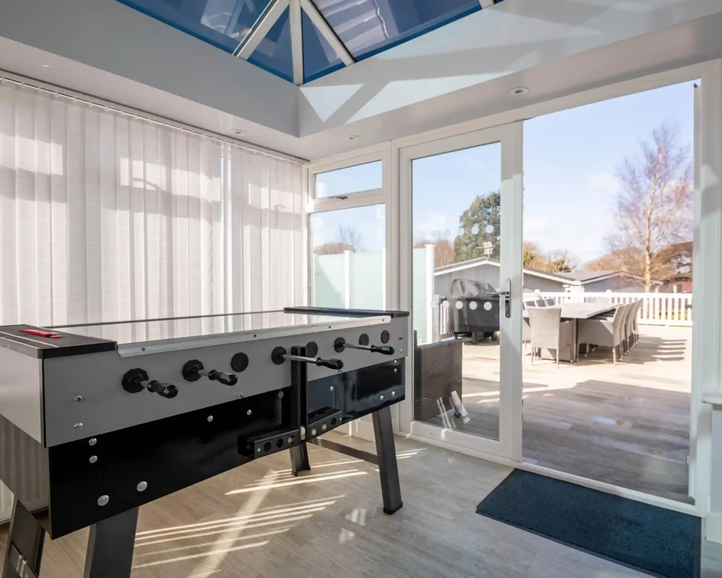 A foosball table is positioned in a bright sunroom with large windows and a glass ceiling. A door leads to an outdoor patio area with seating.