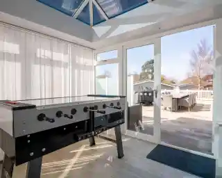 A foosball table is positioned in a bright sunroom with large windows and a glass ceiling. A door leads to an outdoor patio area with seating.