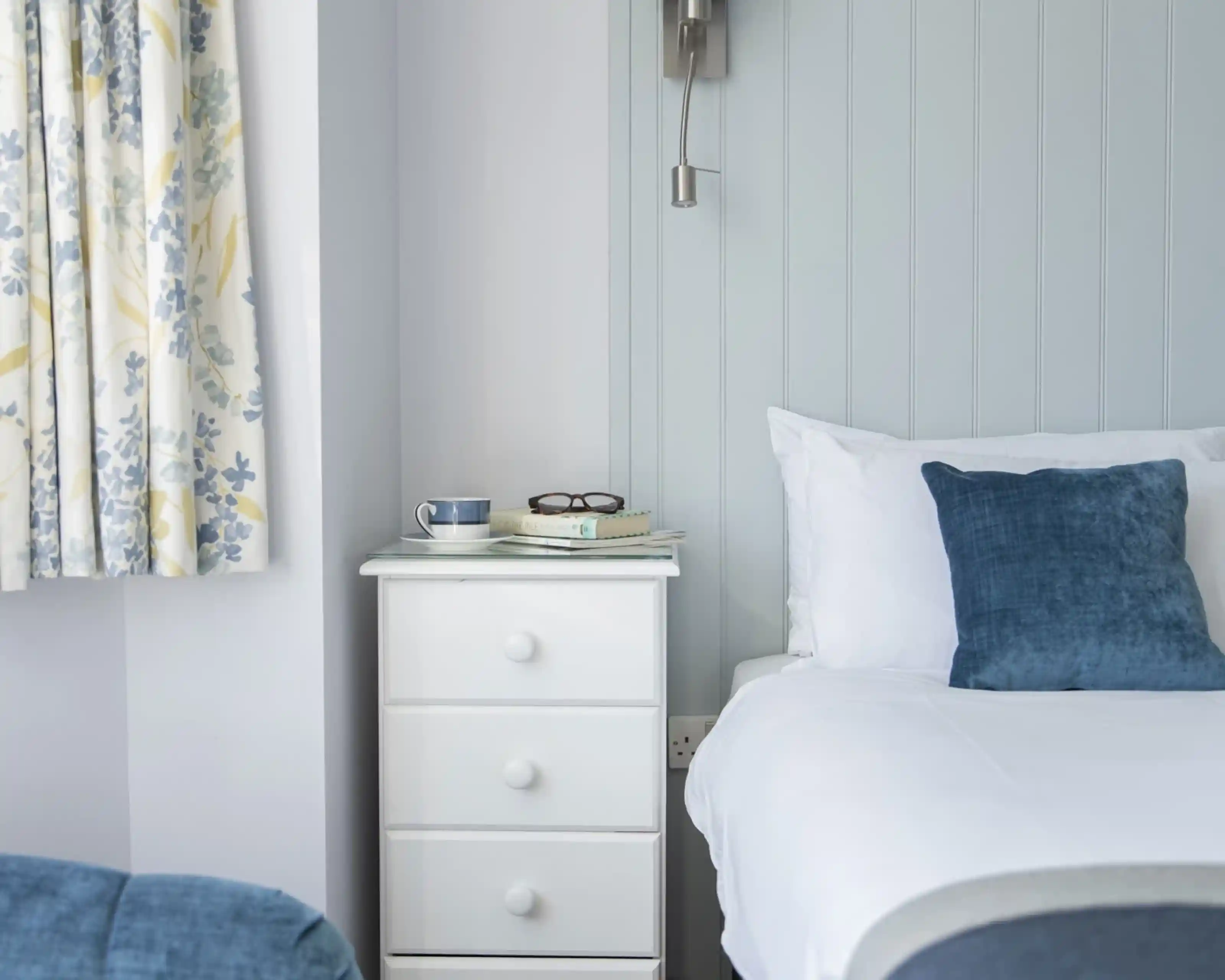 A cozy bedroom featuring a white bedside table with books and glasses, a blue cushion on the bed, and floral curtains in blue and yellow.