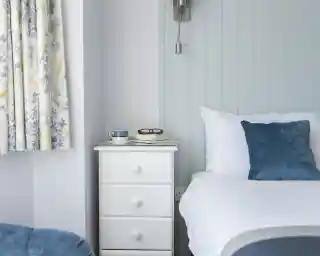 A cozy bedroom featuring a white bedside table with books and glasses, a blue cushion on the bed, and floral curtains in blue and yellow.