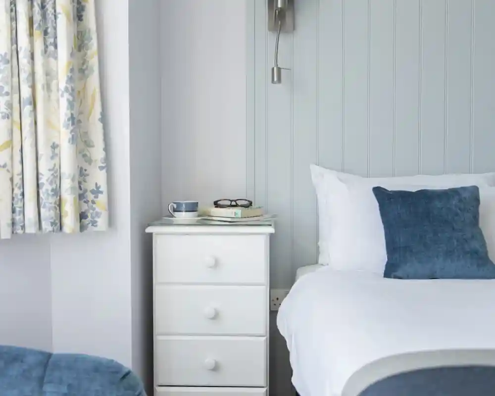 A cozy bedroom featuring a white bedside table with books and glasses, a blue cushion on the bed, and floral curtains in blue and yellow.