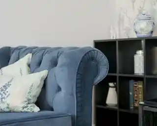 A blue velvet chesterfield sofa with decorative pillows is positioned next to a modern black bookshelf. The bookshelf contains various items, including a vase and books, against a soft-colored wall with floral wallpaper.