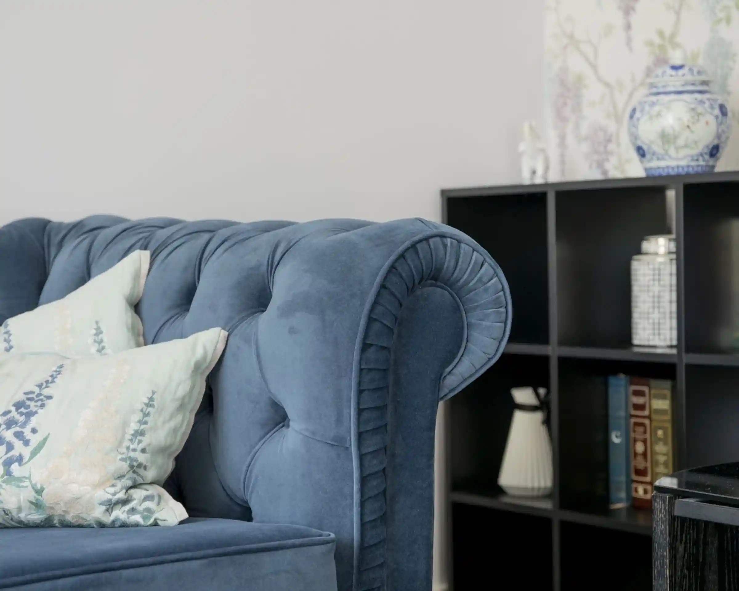 A blue velvet chesterfield sofa with decorative pillows is positioned next to a modern black bookshelf. The bookshelf contains various items, including a vase and books, against a soft-colored wall with floral wallpaper.