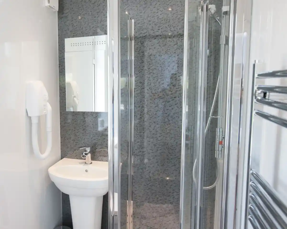 A modern bathroom featuring a glass shower enclosure, a white sink with a chrome faucet, a large mirror, and a sleek towel radiator. The walls have a textured gray finish.
