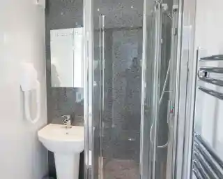 A modern bathroom featuring a glass shower enclosure, a white sink with a chrome faucet, a large mirror, and a sleek towel radiator. The walls have a textured gray finish.