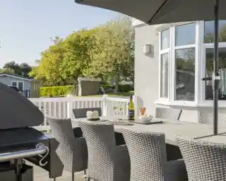 A stylish outdoor dining area with a large table surrounded by wicker chairs. A gas grill is visible in the foreground. The setting features green trees and a bright, sunny atmosphere, suggesting a relaxing environment. A bottle of wine and glasses are placed on the table alongside a plate of snacks.