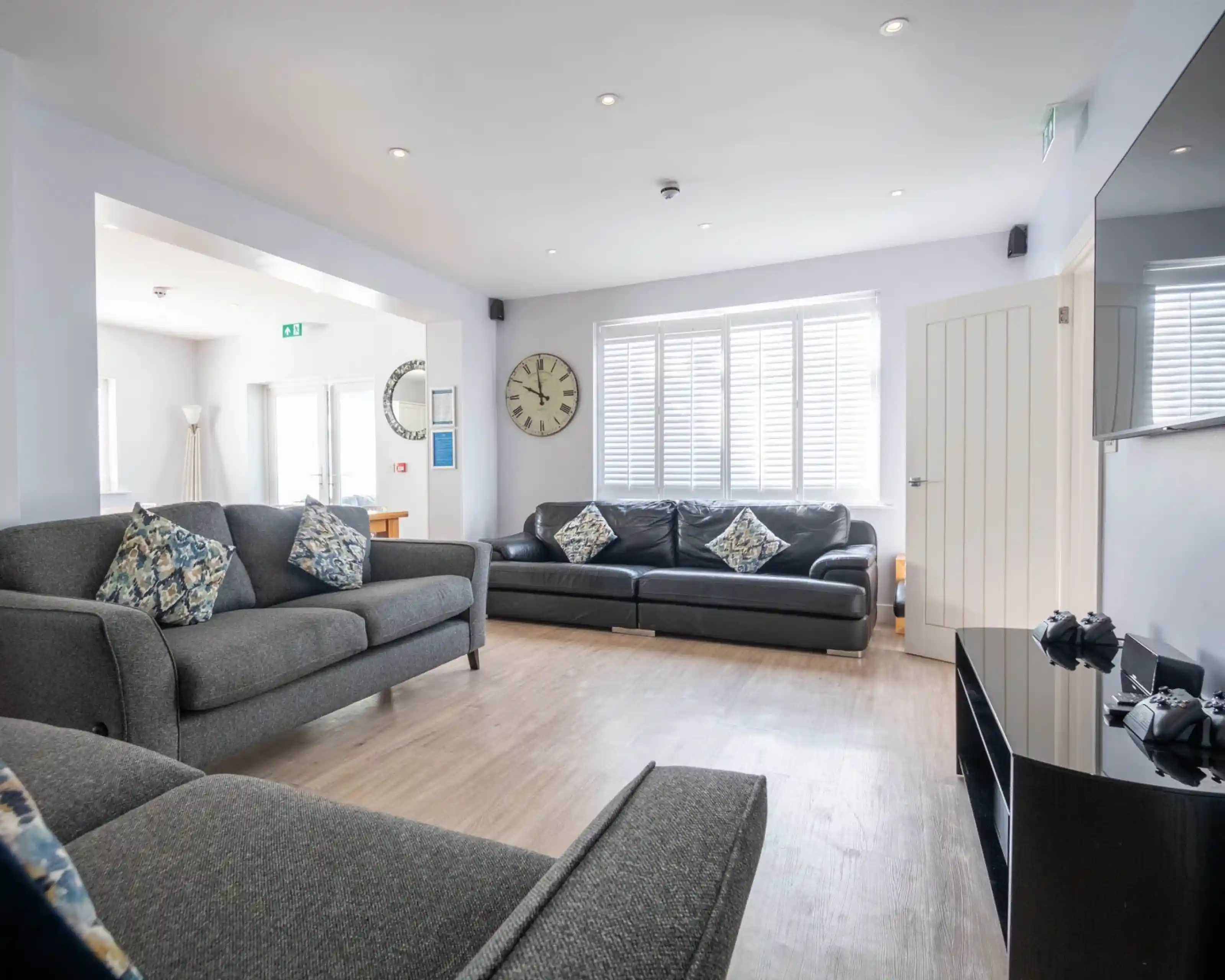 A modern living room featuring two sofas with decorative cushions, a large wall clock, and a flat-screen TV mounted on the wall. Bright natural light streams through the windows.