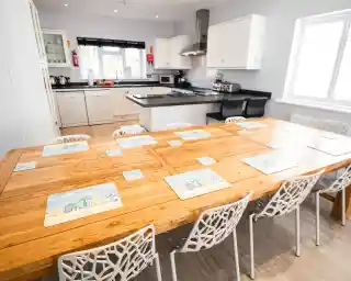 A spacious kitchen with a large wooden dining table set for eight, featuring colorful placemats. Modern appliances and cabinetry are visible in the background. Natural light fills the bright, airy room.