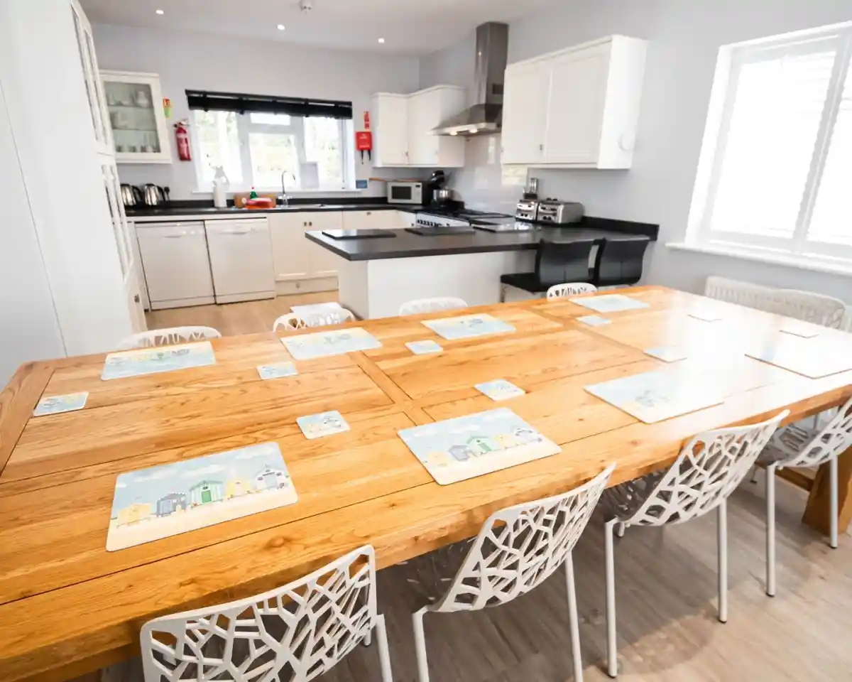 A spacious kitchen with a large wooden dining table set for eight, featuring colorful placemats. Modern appliances and cabinetry are visible in the background. Natural light fills the bright, airy room.