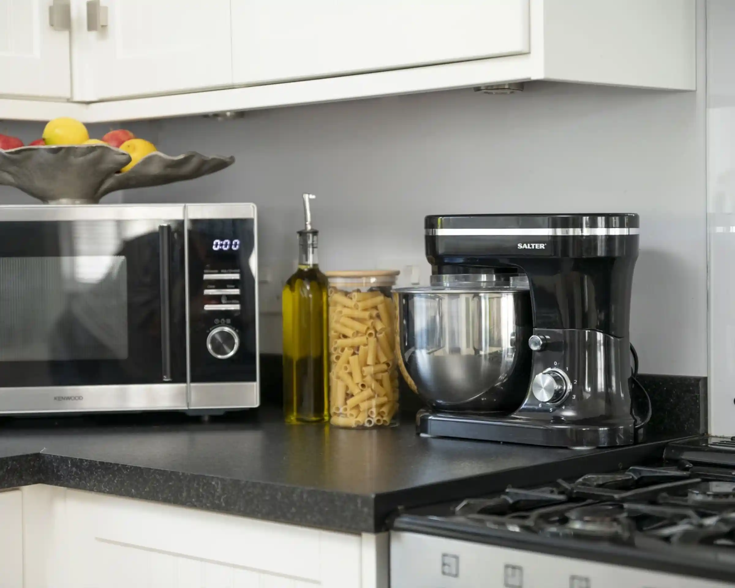 A modern kitchen countertop featuring a silver microwave, a black stand mixer, a glass jar filled with pasta, an olive oil bottle, and a decorative bowl of fruits.