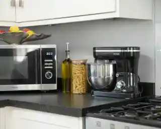 A modern kitchen countertop featuring a silver microwave, a black stand mixer, a glass jar filled with pasta, an olive oil bottle, and a decorative bowl of fruits.