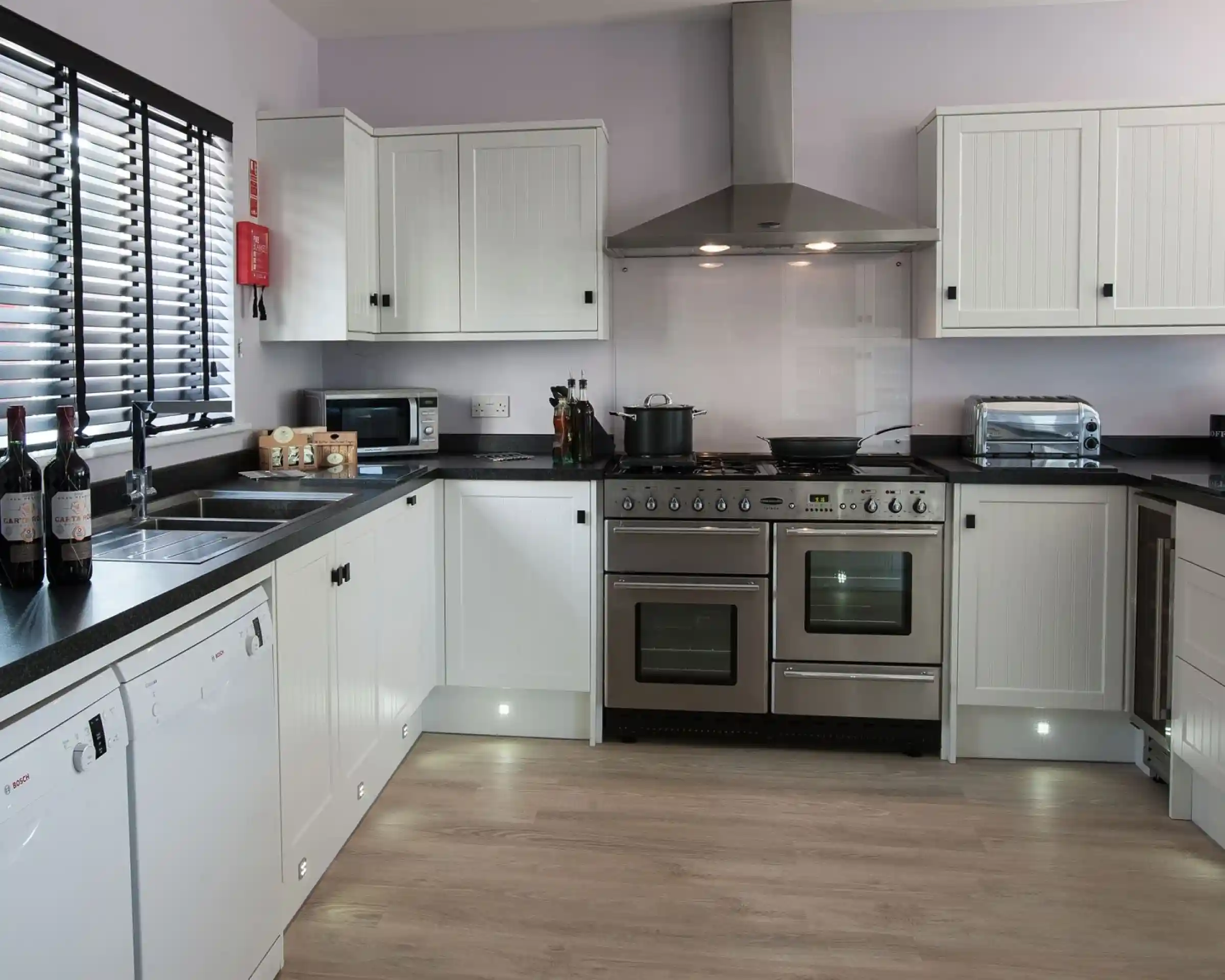 Modern kitchen featuring white cabinetry, stainless steel appliances, and a black countertop. The space includes a stove, microwave, and wine bottles on the counter, with light wood flooring and large windows for natural light.