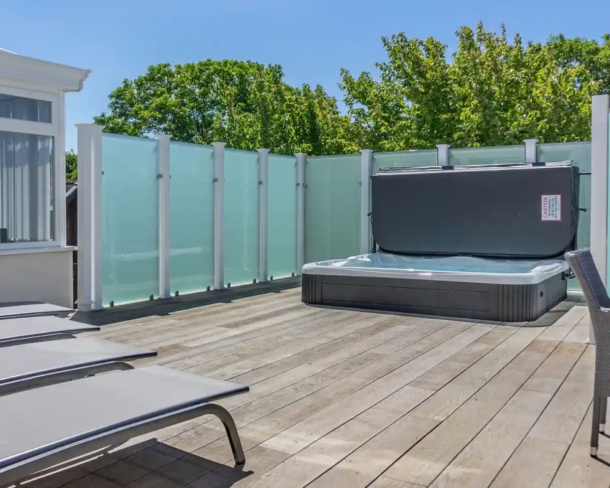 A wooden deck featuring a hot tub, surrounded by clear glass panels for privacy. Sun loungers are positioned nearby, and lush green trees are visible in the background under a blue sky.