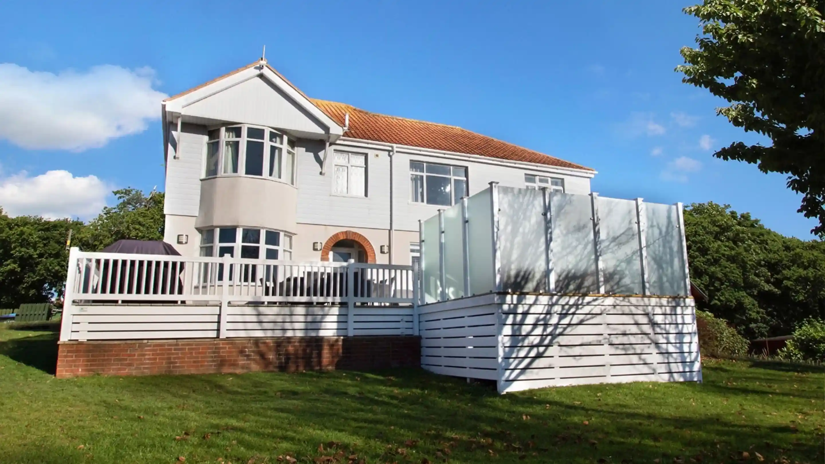 A large, modern two-story house with a light gray exterior and orange roof tiles. The house features large windows and a white wooden deck. In front, there is a terrace with a glass panel enclosure. Lush green grass surrounds the property under a clear blue sky.