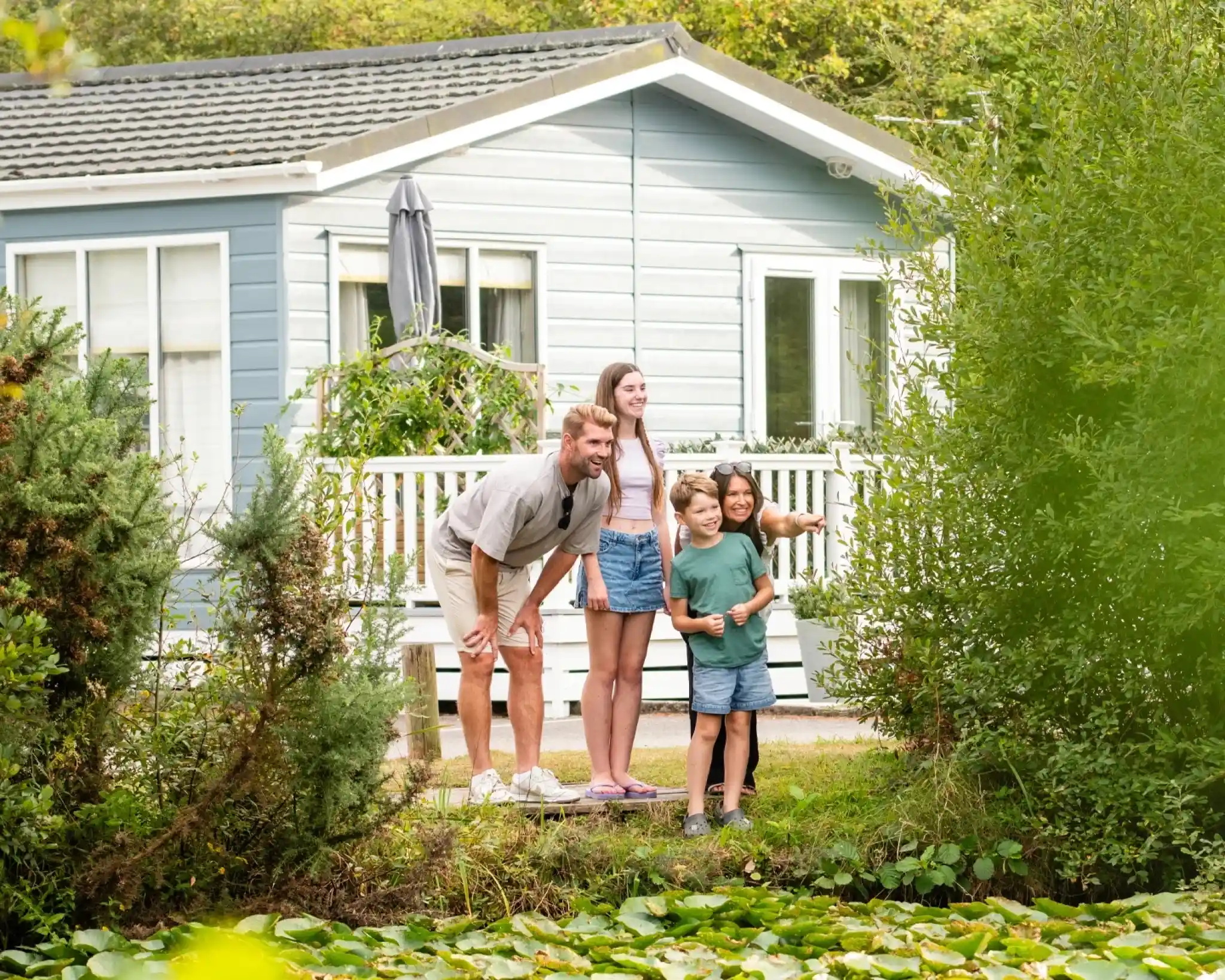 A family of four stands on a wooden platform near a pond, smiling and looking out towards the water. A light blue house is visible in the background, surrounded by greenery and trees.
