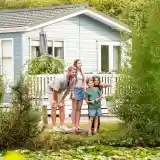 A family of four stands on a wooden platform near a pond, smiling and looking out towards the water. A light blue house is visible in the background, surrounded by greenery and trees.