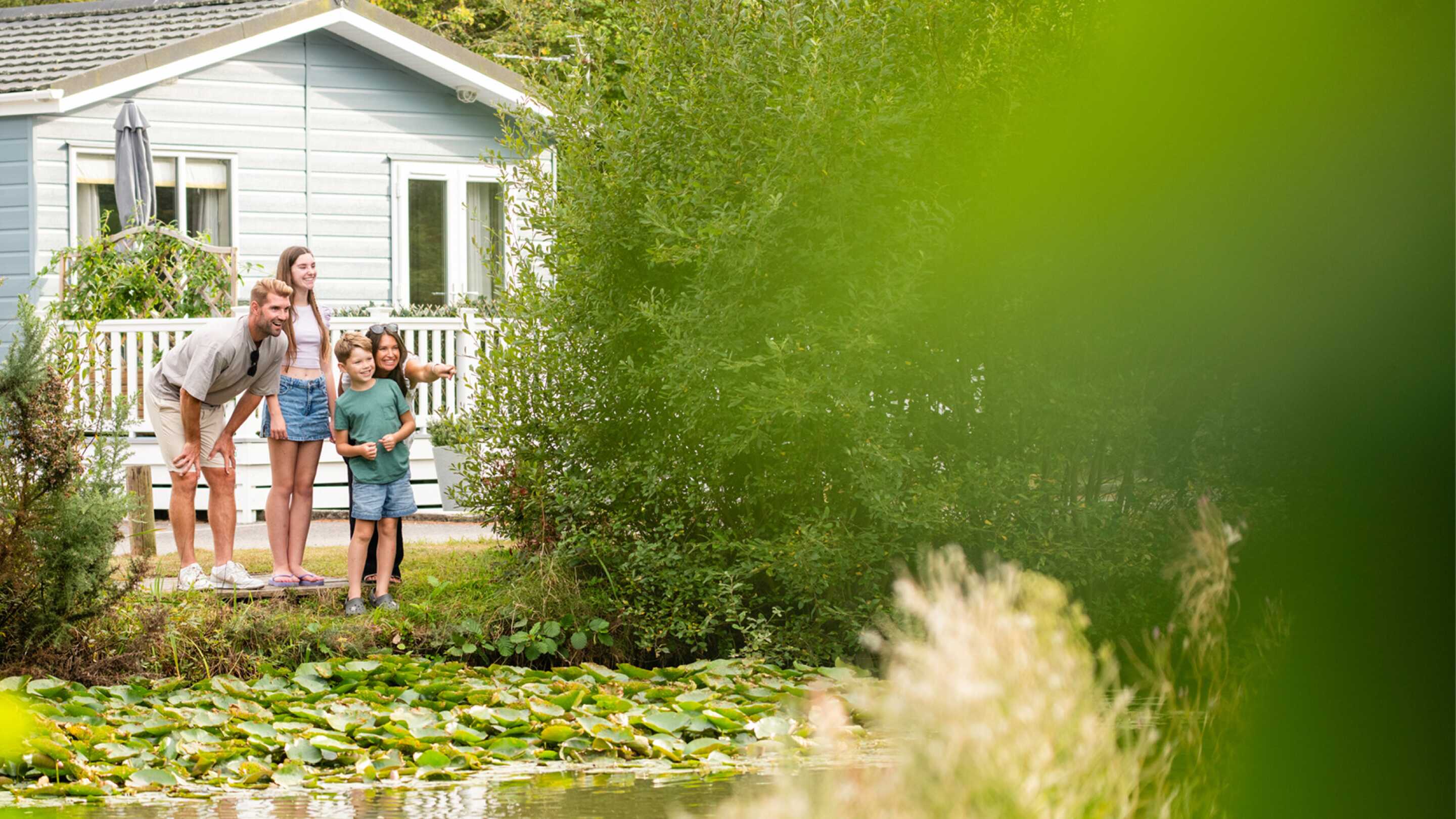 A woman, a girl, and a boy stand by a pond surrounded by lush greenery, smiling as they look at something in the water. A blue cottage is visible in the background, partially obscured by trees.