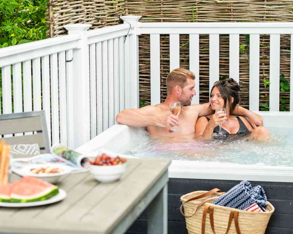 A couple shares a joyful moment in a hot tub, holding glasses of sparkling drinks. They are seated closely, smiling at each other. In the foreground, a table is set with a plate of snacks, including fruits and crackers, and a beach bag is nearby. Lush greenery surrounds the scene, adding to the relaxed atmosphere.