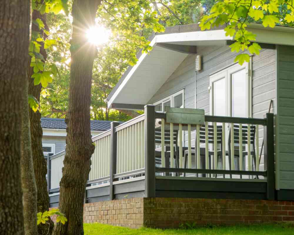 A modern cabin with a spacious deck is set among trees, illuminated by sunlight filtering through the leaves. The cabin features large windows and a sloped roof, creating a serene outdoor atmosphere.