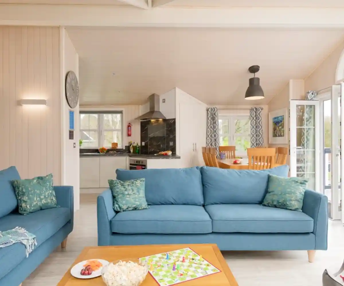 A row of static caravans is set in a lush green area, with a circular flower bed filled with vibrant blooms in the foreground. Tall trees and a clear blue sky complete the tranquil scene.
