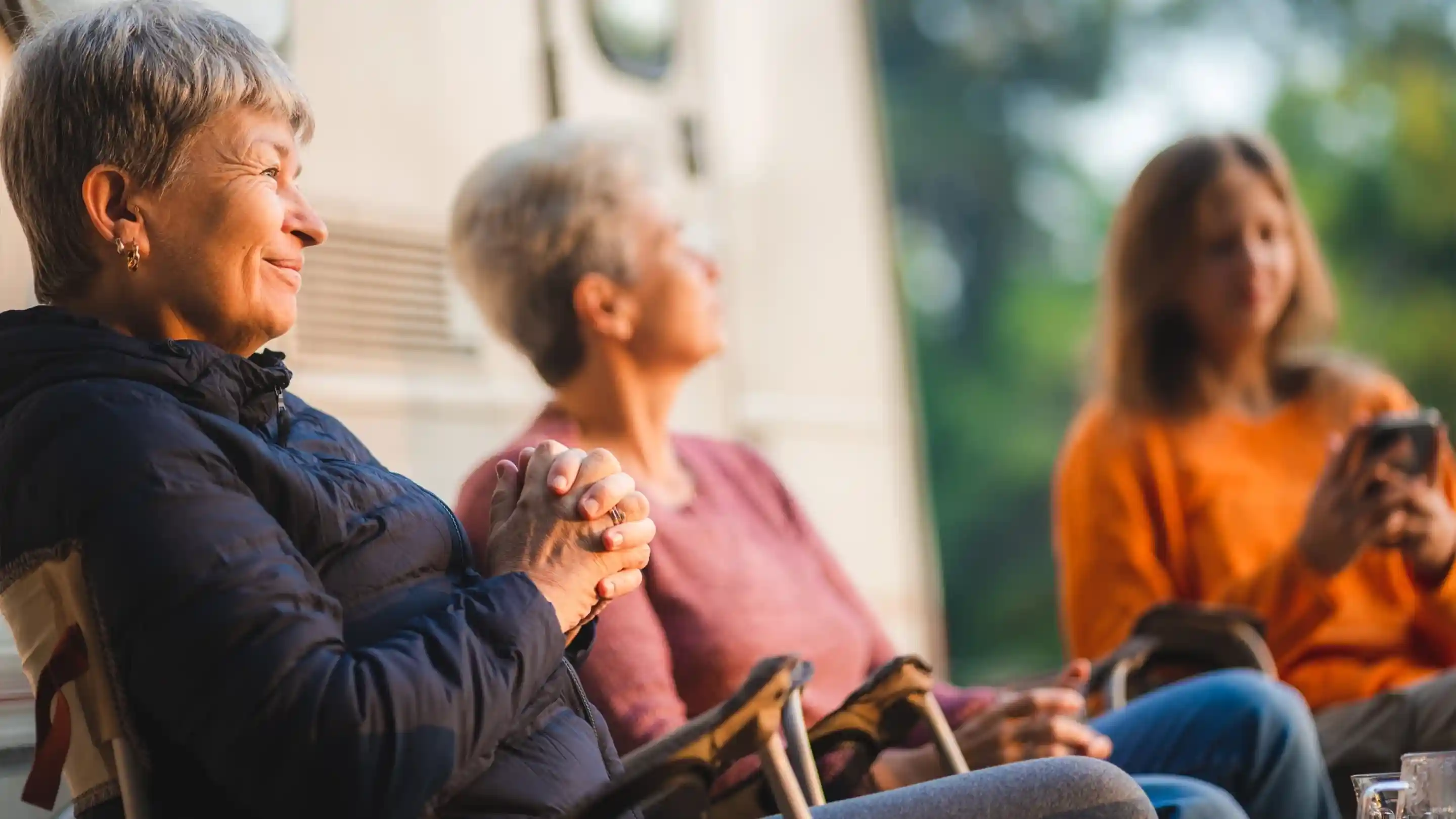 Three women enjoying a relaxed moment outdoors, sitting in camping chairs. One woman smiles warmly, while another looks off to the side. A third woman is engaged with her phone, all surrounded by nature.