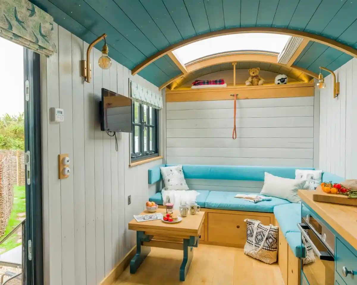 A cozy tiny home interior featuring a light blue sectional couch, wooden furniture, and a small TV on the wall. Natural light streams through a large window, illuminating the space decorated with soft cushions and a small shelf with a teddy bear. A table holds snacks and drinks, adding to the inviting atmosphere.