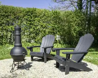 Two black Adirondack chairs sit beside a round outdoor fire pit in a garden, surrounded by greenery and bright blue sky.