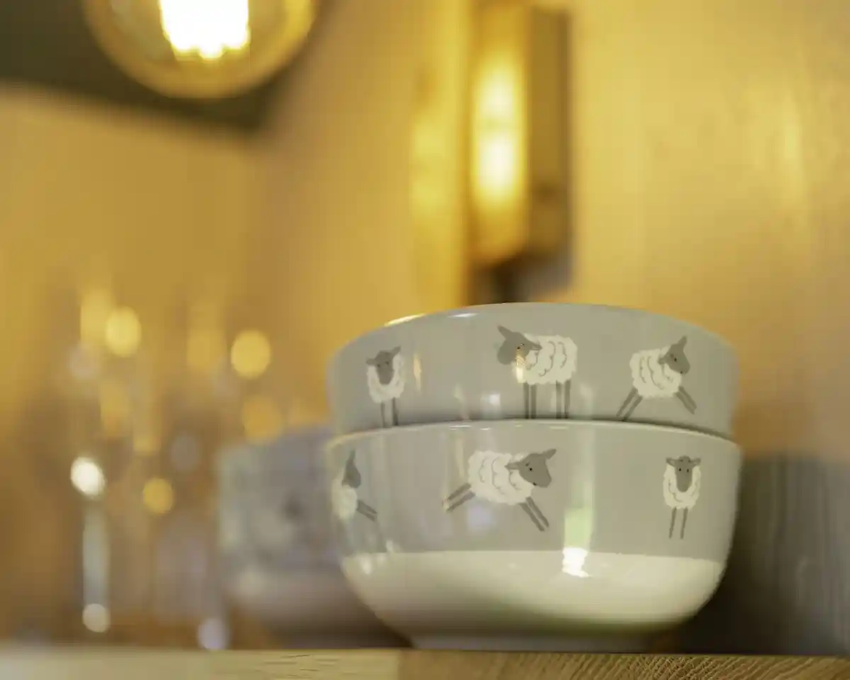 Stacked bowls with sheep designs rest on a wooden shelf, illuminated by warm lighting. Blurry glassware is visible in the background.