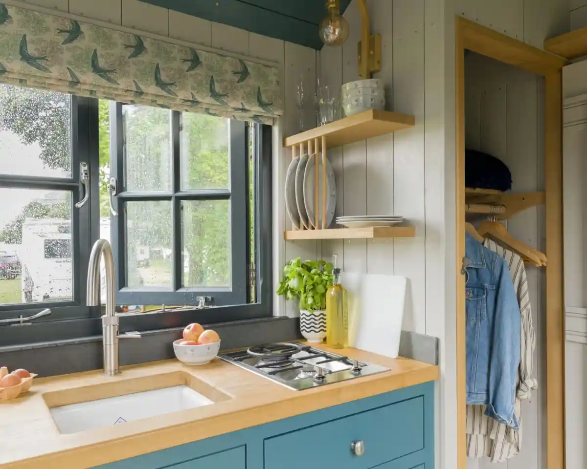 A cozy kitchen with a blue cabinet featuring a wooden countertop and a white sink. A modern faucet and a gas stove are visible. Behind the sink, a window with curtains overlooks greenery, and shelves hold plates and a plant. A hanging area on the right displays a denim jacket.