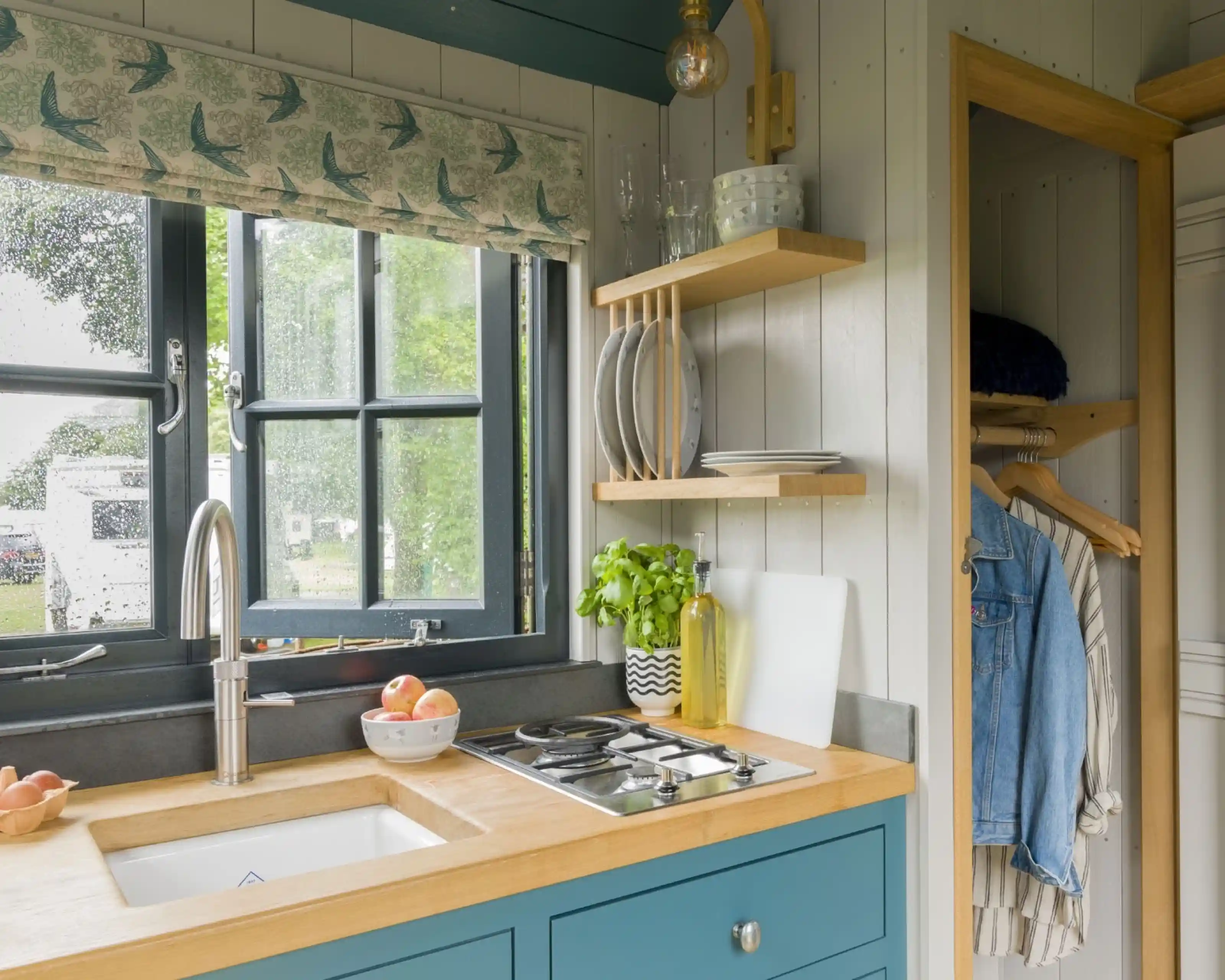 A cozy kitchen with a blue cabinet featuring a wooden countertop and a white sink. A modern faucet and a gas stove are visible. Behind the sink, a window with curtains overlooks greenery, and shelves hold plates and a plant. A hanging area on the right displays a denim jacket.