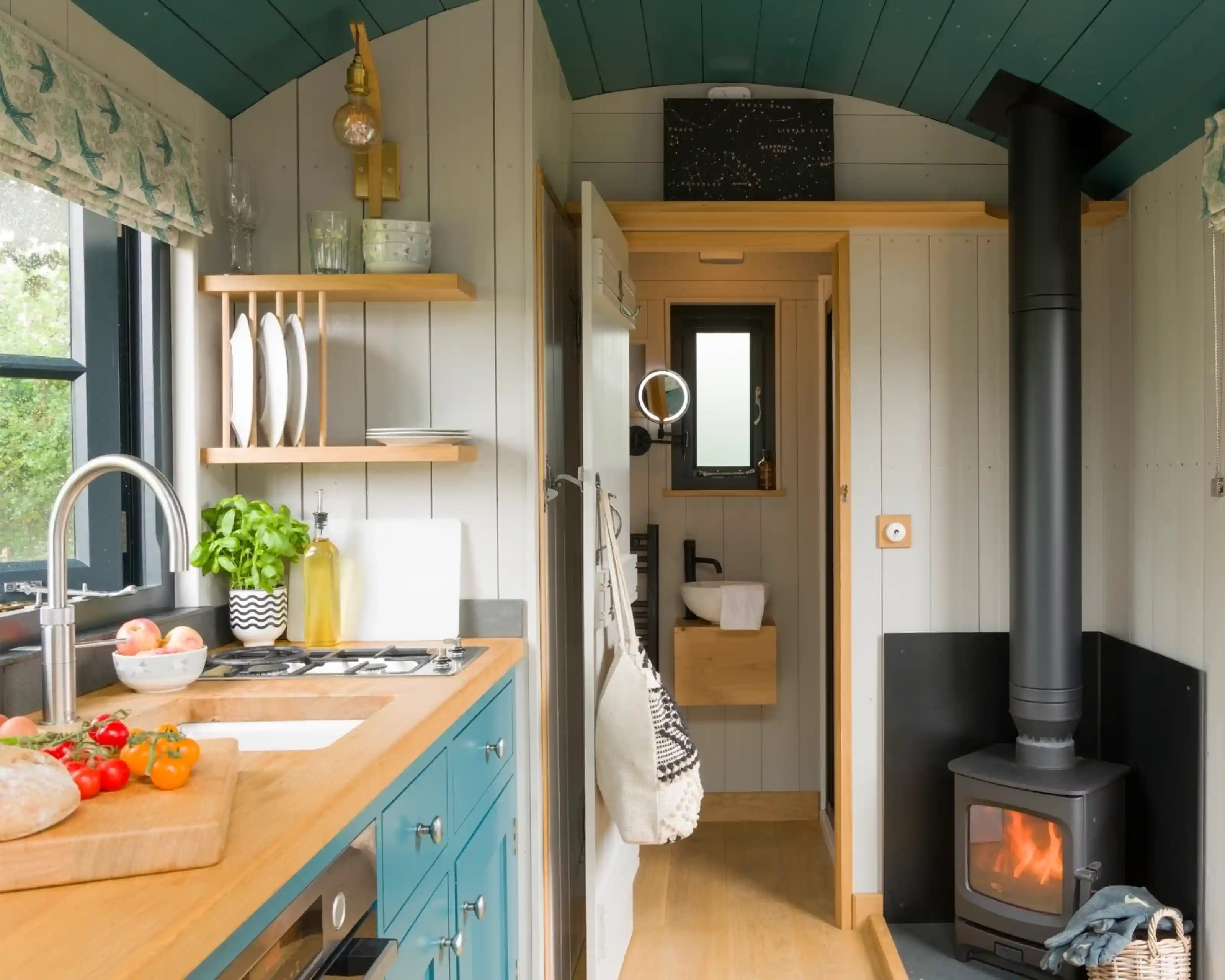 Cozy tiny kitchen with a blue and white color scheme, featuring a wooden countertop, a stove, and a small wood-burning stove. A plant sits near the sink, and there are plates displayed on a shelf. A doorway leads to a bathroom area with modern fixtures. Natural light comes through a window.
