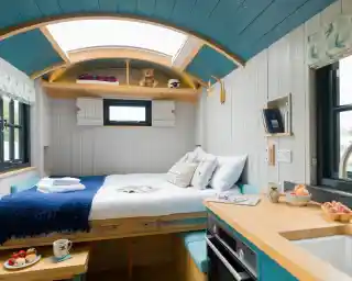 A cozy tiny home interior featuring a double bed with white linens and decorative pillows. The walls are painted white with blue accents, and there are large windows allowing for natural light. A small kitchen area includes a sink and a few snacks on the counter. A teddy bear and decorative items are placed on shelves above the bed.