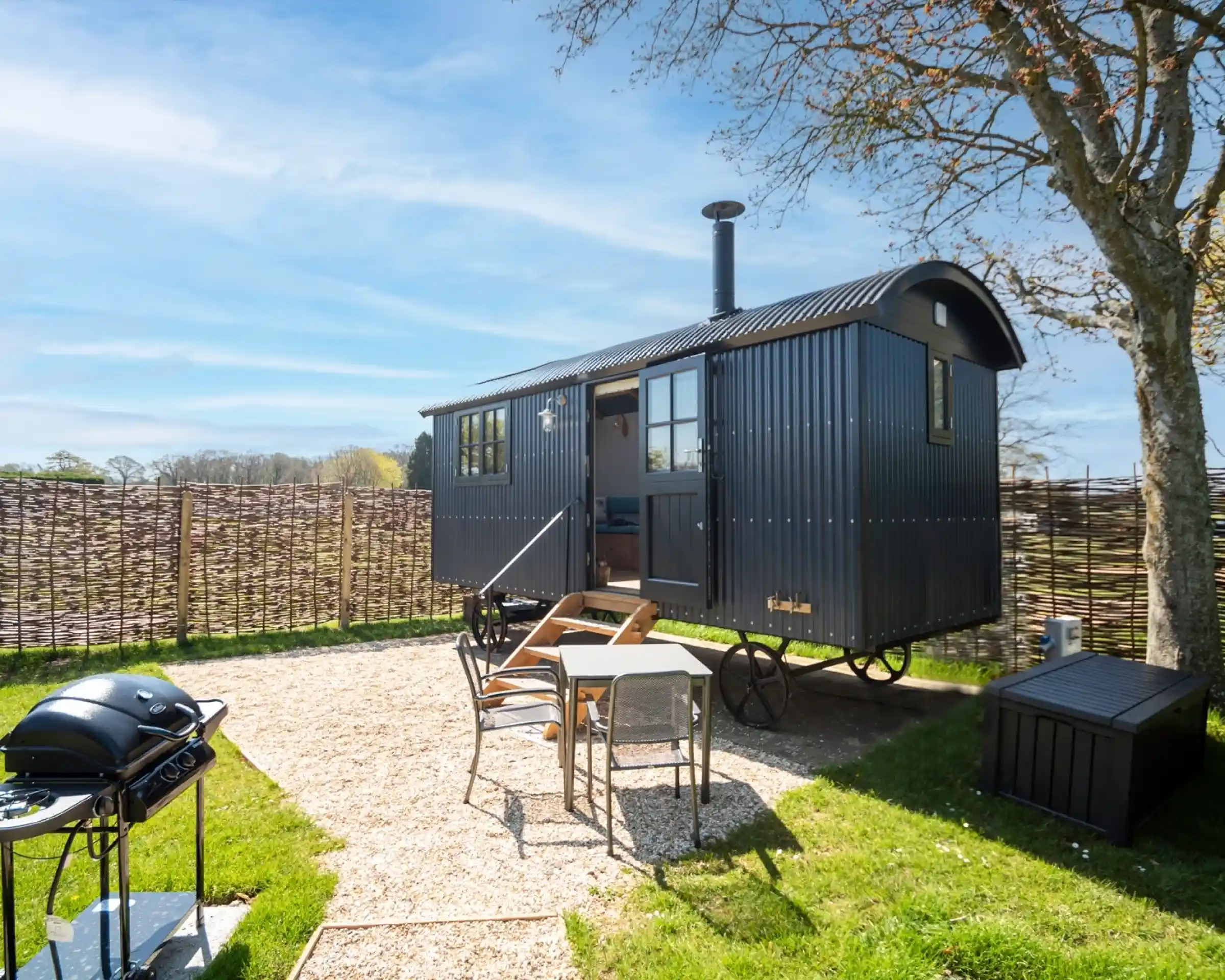 A black, vintage-style caravan is set on a gravel patch surrounded by green grass. There are wooden steps leading to the entrance, a BBQ grill nearby, and a small table with chairs under a tree. The sky is clear with a few clouds.