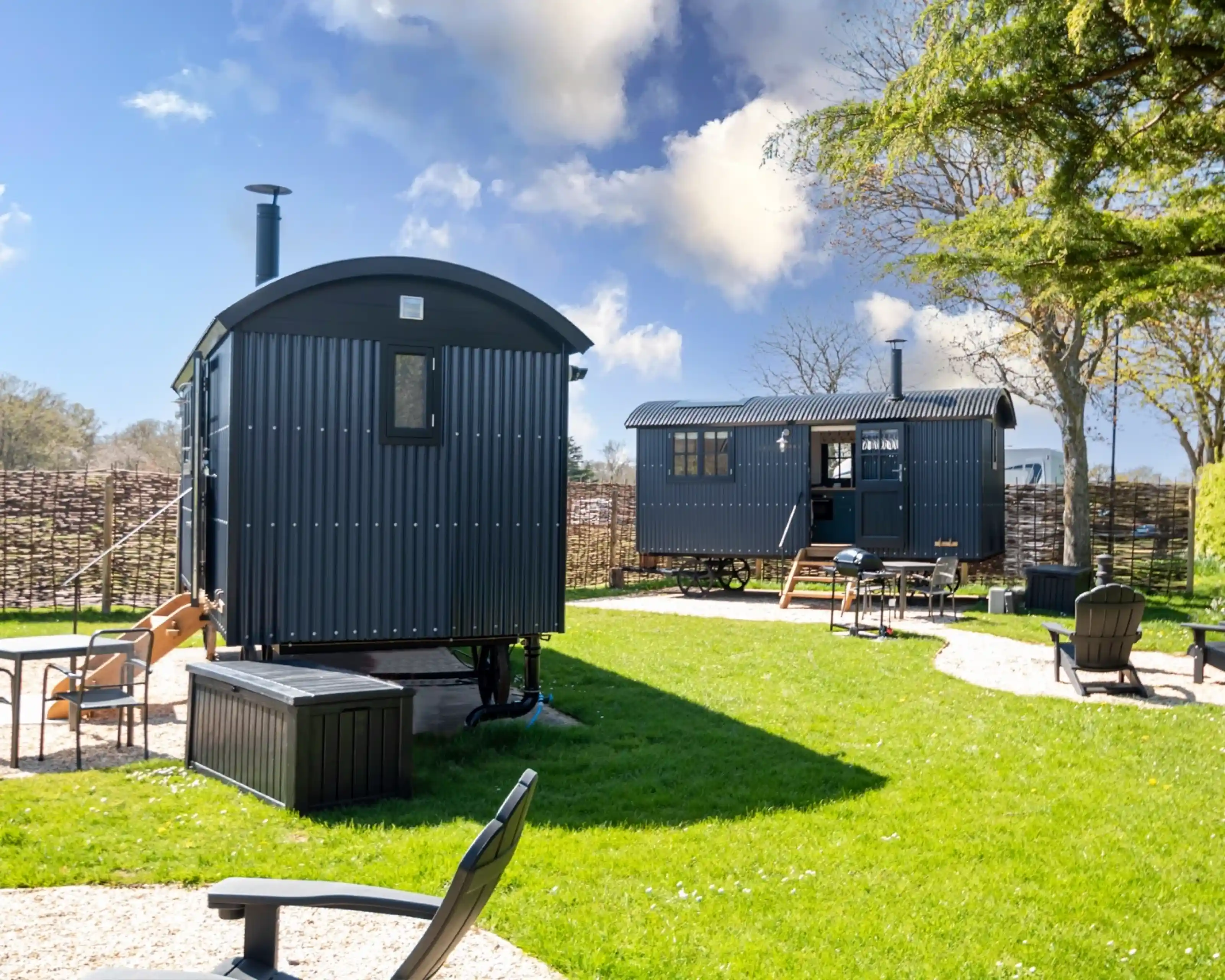 Two stylish shepherd huts sit in a grassy yard, with a clear blue sky and fluffy clouds above. One hut is slightly smaller, featuring a wooden deck and a slide, while the larger hut has a chimney and a porch area. Surrounding the huts are circular stone paths and outdoor seating in the form of black chairs.
