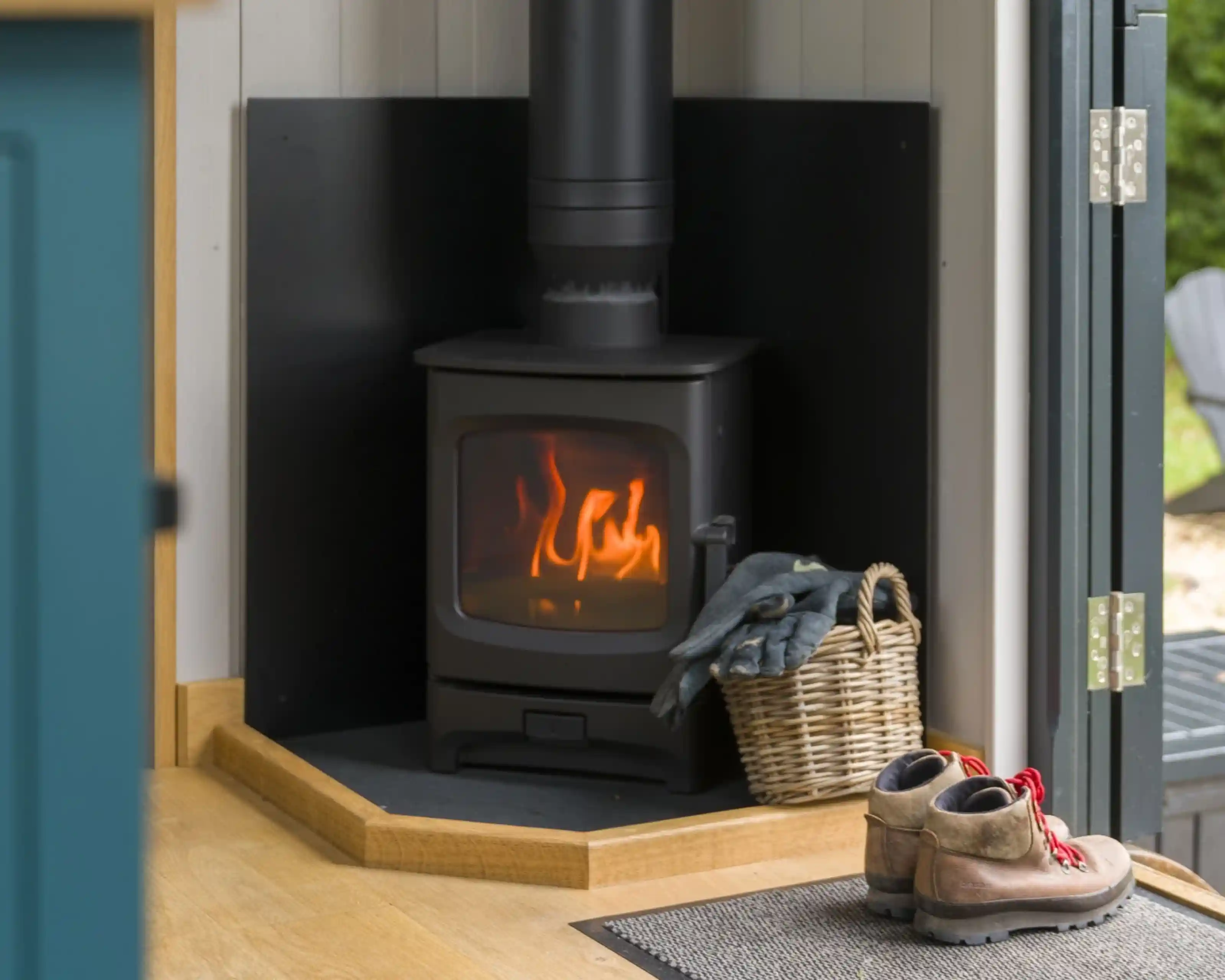 A modern wood stove emits a warm glow, situated in a corner with black backing. Nearby, a woven basket holds blankets, and a pair of rugged boots rests on a mat by the door.